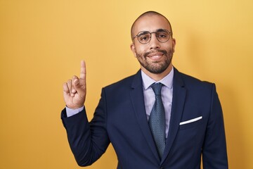 Hispanic man with beard wearing suit and tie showing and pointing up with finger number one while smiling confident and happy.