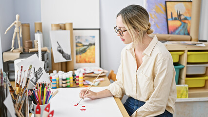 Young blonde woman artist drawing on paper sitting on table at art studio