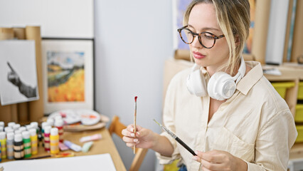 Young blonde woman artist sitting on table choosing paintbrush wearing headphones at art studio