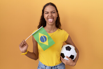 Young hispanic woman holding brazil flag and football ball sticking tongue out happy with funny...