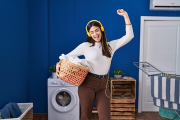 Young beautiful hispanic woman listening to music holding basket with clothes at laundry room