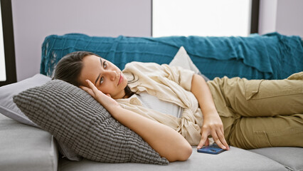 Unhappy young hispanic woman lying on the sofa at home, deeply engrossed in a serious smartphone...