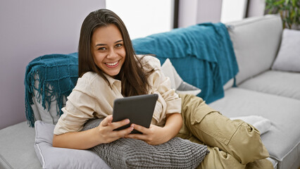 Irresistibly confident young beautiful hispanic woman joyfully using touchpad while relaxing on her comfy sofa, exuding positivity in the cozy ambiance of her home living room.
