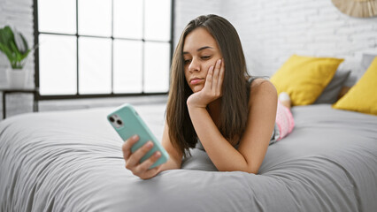 Upset young hispanic woman lying comfortably in bed using her smartphone. beautiful female relaxing...