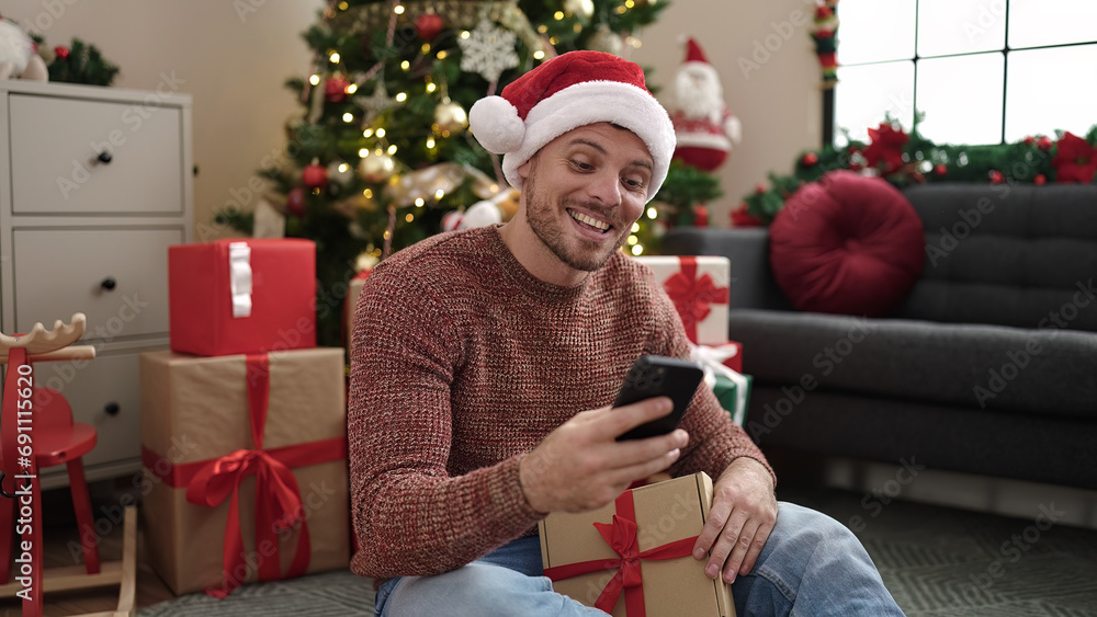 Poster young caucasian man using smartphone sitting on the floor by christmas tree at home