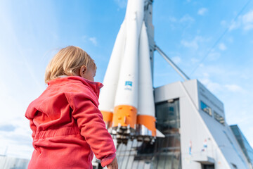 A cute child toddler girl stands in front of the building of the Space Museum and Exhibition Center in Samara, Russia, against the background of the Soyuz launch vehicle. Samara, Russia - 30 Sep 2023