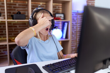 Young caucasian woman playing video games wearing headphones peeking in shock covering face and eyes with hand, looking through fingers with embarrassed expression.