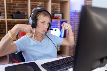 Young caucasian woman playing video games wearing headphones pointing down looking sad and upset,...