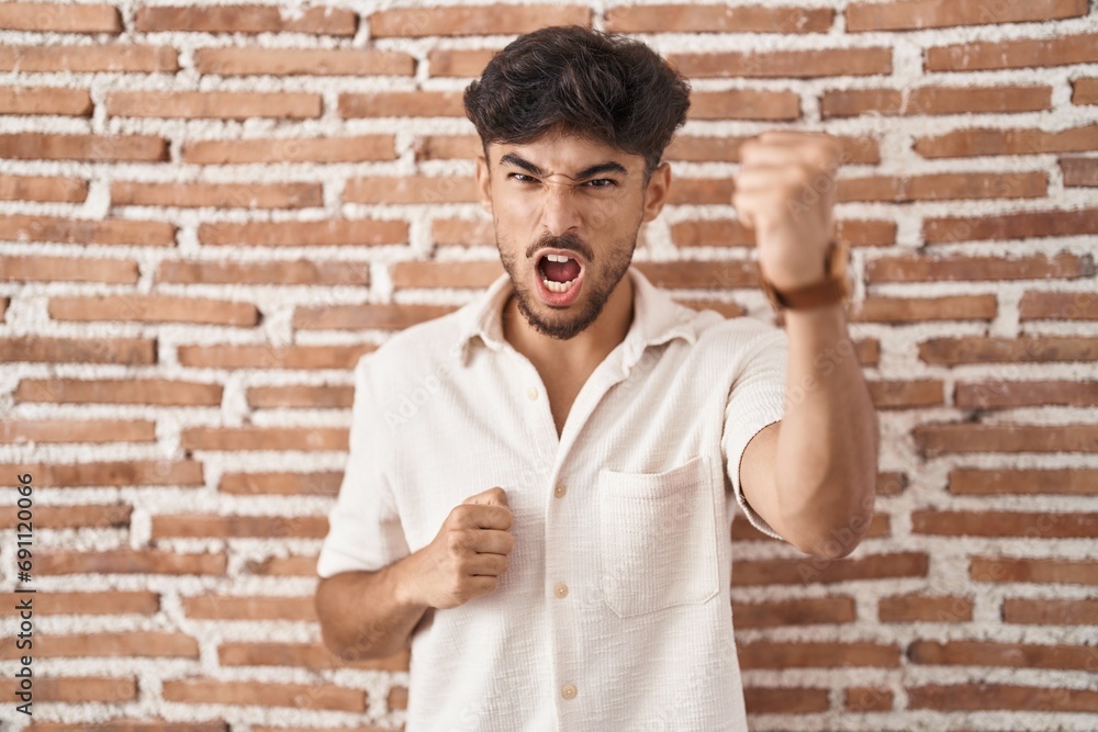 Wall mural Arab man with beard standing over bricks wall background angry and mad raising fist frustrated and furious while shouting with anger. rage and aggressive concept.