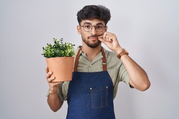 Arab man with beard holding green plant pot mouth and lips shut as zip with fingers. secret and...