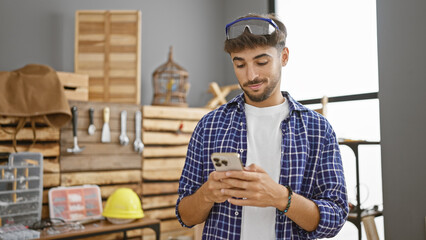 Smiling young arab man, a professional carpenter, cheerfully texts on his smartphone amidst buzzing...