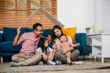 A joyful family with kids mother father sits on a sofa holding a cardboard roof symbolizing...