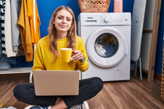 Young blonde woman using laptop drinking coffee waiting for washing machine at laundry room