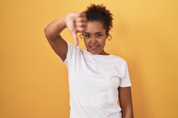 Young hispanic woman with curly hair standing over yellow background looking unhappy and angry showing rejection and negative with thumbs down gesture. bad expression.