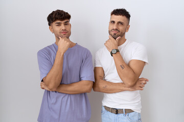 Homosexual gay couple standing over white background looking confident at the camera smiling with crossed arms and hand raised on chin. thinking positive.