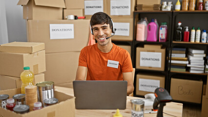 Handsome young hispanic man selflessly volunteering, working on laptop with headphones at charity...