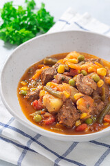 Beef stew with potato, green beans, carrot, peas and corn, in gray bowl, vertical