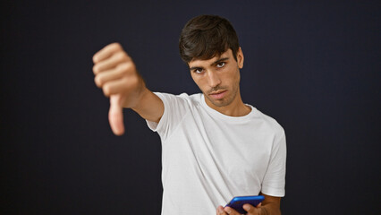 Cool, young hispanic man in casual fashion delivering a negative thumb down gesture, looking unhappily at his smartphone, expressing failure, standing against an isolated black background.