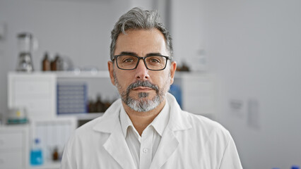 Handsome, grey-haired young hispanic male scientist seriously focused on medical experiment indoors in a vibrant lab - concentrating on analysis with microscope while standing at the work table - obrazy, fototapety, plakaty