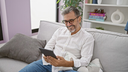 Confident young hispanic grey-haired man enjoys using touchpad while comfortably sitting on sofa...