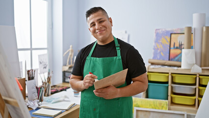 Confident young latin artist, apron-clad and smiling, masterfully wielding his paintbrush and palette indoors at his lively art studio