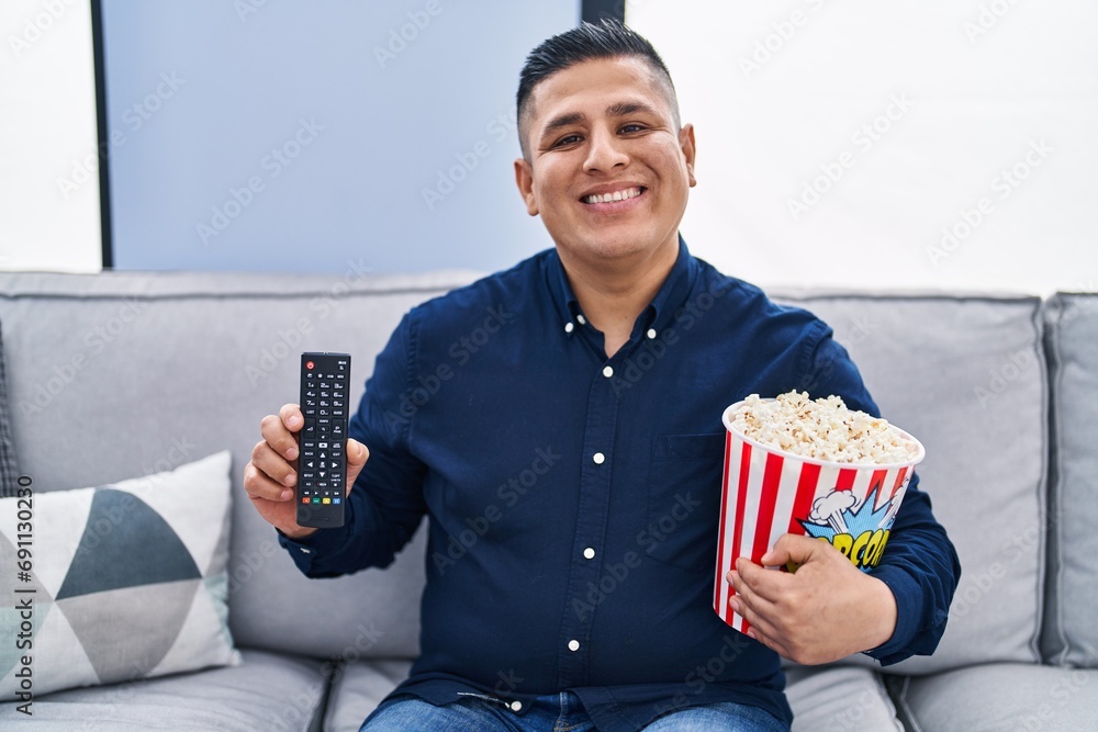 Poster hispanic young man eating popcorn using tv control smiling with a happy and cool smile on face. show