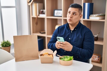 Hispanic young man eating take away food using smartphone smiling looking to the side and staring...