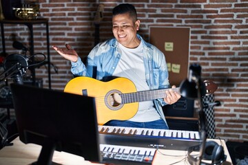 Hispanic young man playing classic guitar at music studio smiling cheerful presenting and pointing with palm of hand looking at the camera.