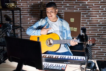 Hispanic young man playing classic guitar at music studio pointing aside worried and nervous with...