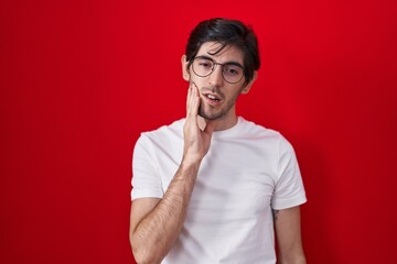 Young hispanic man standing over red background touching mouth with hand with painful expression...