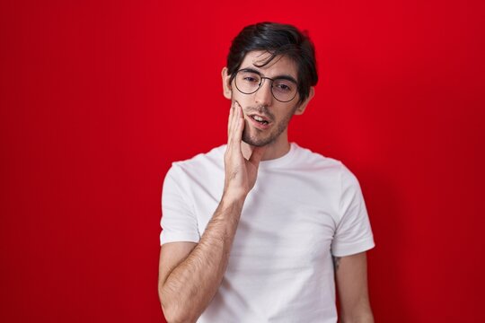 Young Hispanic Man Standing Over Red Background Touching Mouth With Hand With Painful Expression Because Of Toothache Or Dental Illness On Teeth. Dentist