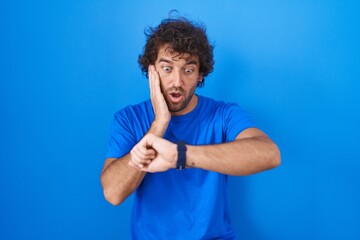 Hispanic young man standing over blue background looking at the watch time worried, afraid of getting late