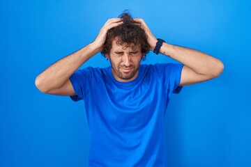 Hispanic young man standing over blue background suffering from headache desperate and stressed because pain and migraine. hands on head.