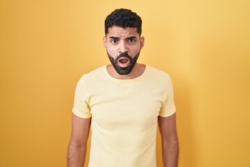 Hispanic man with beard standing over yellow background in shock face, looking skeptical and sarcastic, surprised with open mouth