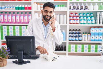 Young arab man pharmacist talking on telephone using computer at pharmacy