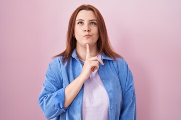 Young hispanic woman with red hair standing over pink background thinking concentrated about doubt...