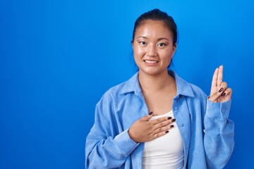 Asian young woman standing over blue background smiling swearing with hand on chest and fingers up, making a loyalty promise oath