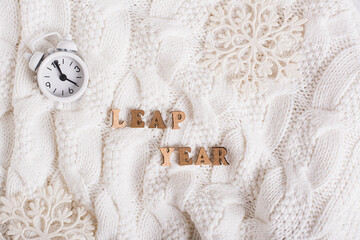 Leap year concept, text made of wooden letters, alarm clock and snowflakes on a knitted background
