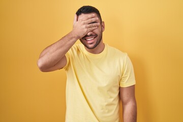 Young hispanic man standing over yellow background smiling and laughing with hand on face covering...