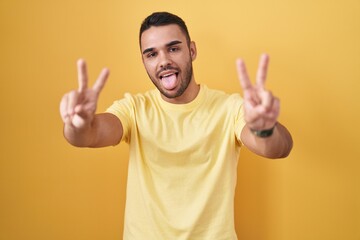 Young hispanic man standing over yellow background smiling with tongue out showing fingers of both hands doing victory sign. number two.