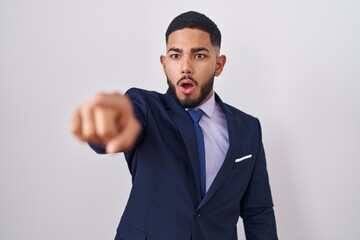 Young hispanic man wearing business suit and tie pointing with finger surprised ahead, open mouth amazed expression, something on the front