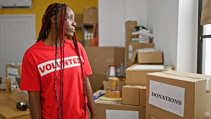 Beautiful african american woman, a serious-faced volunteer, stands indoors in charity center's...