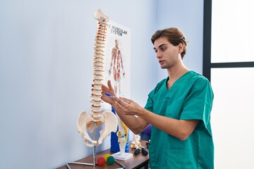 Young caucasian man physiotherapist touching anatomical model of spinal column at rehab clinic