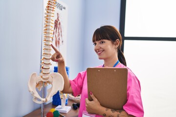 Young beautiful hispanic woman physiotherapist pointing to anatomical model of spinal column...