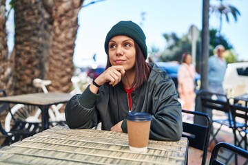 Young hispanic woman drinking a cup of coffee outdoors serious face thinking about question with...