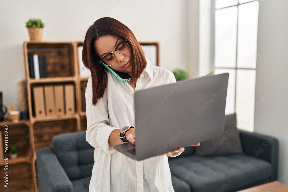 Wall mural young beautiful hispanic woman business worker talking on smartphone using laptop at office