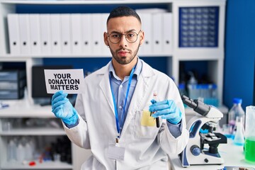 Young hispanic man working at scientist laboratory holding your donation matters holding blood...