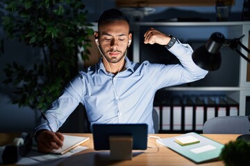 Young hispanic man working at the office at night strong person showing arm muscle, confident and...