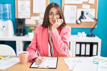 Young hispanic woman working at the office wearing glasses looking stressed and nervous with hands...