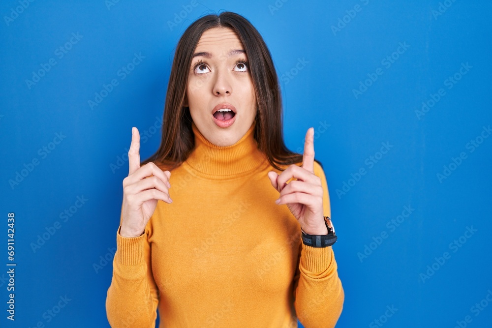 Wall mural young brunette woman standing over blue background amazed and surprised looking up and pointing with